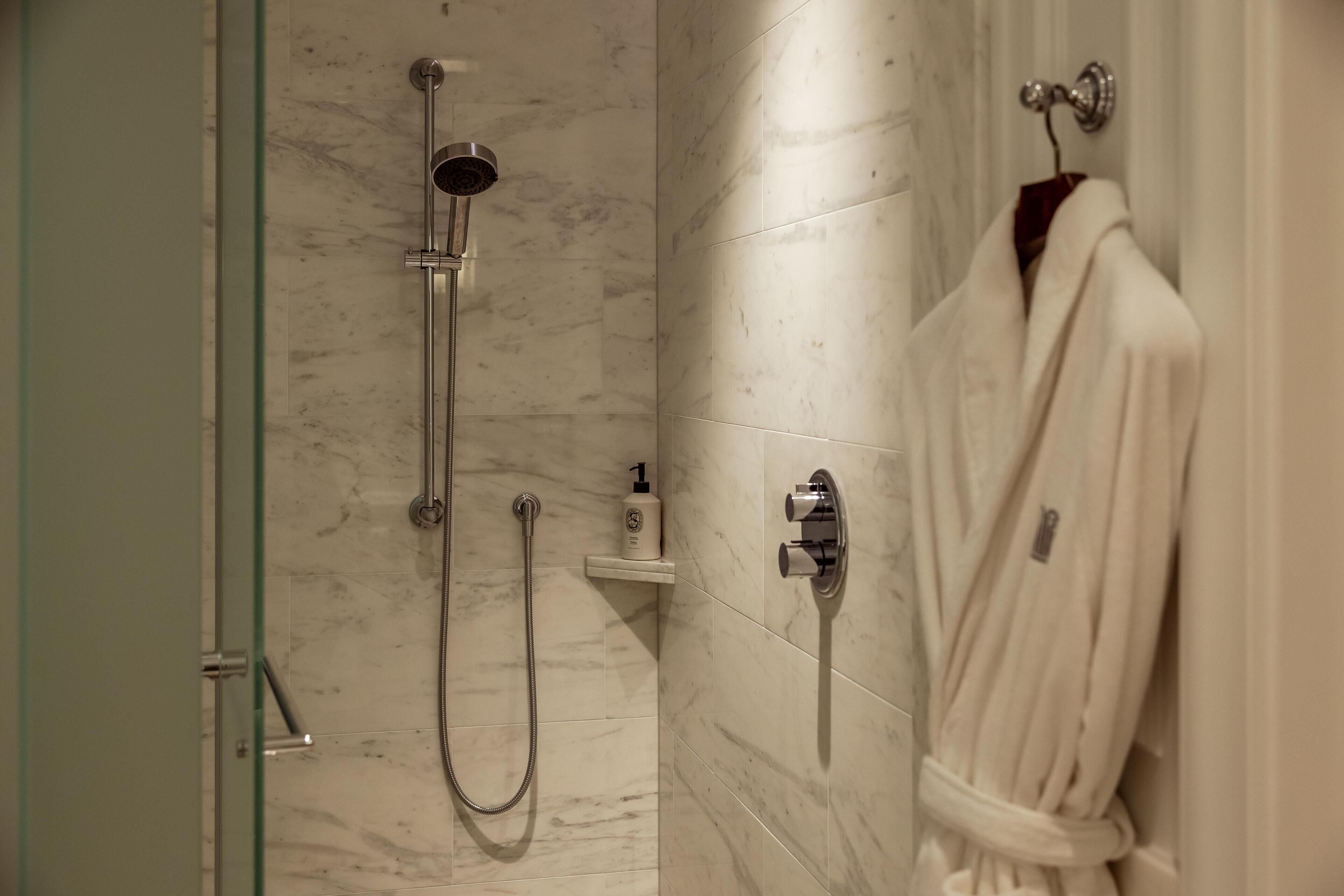 A bathroom with a glass shower enclosure, a white robe hanging on a hook, and a small shelf with toiletries.