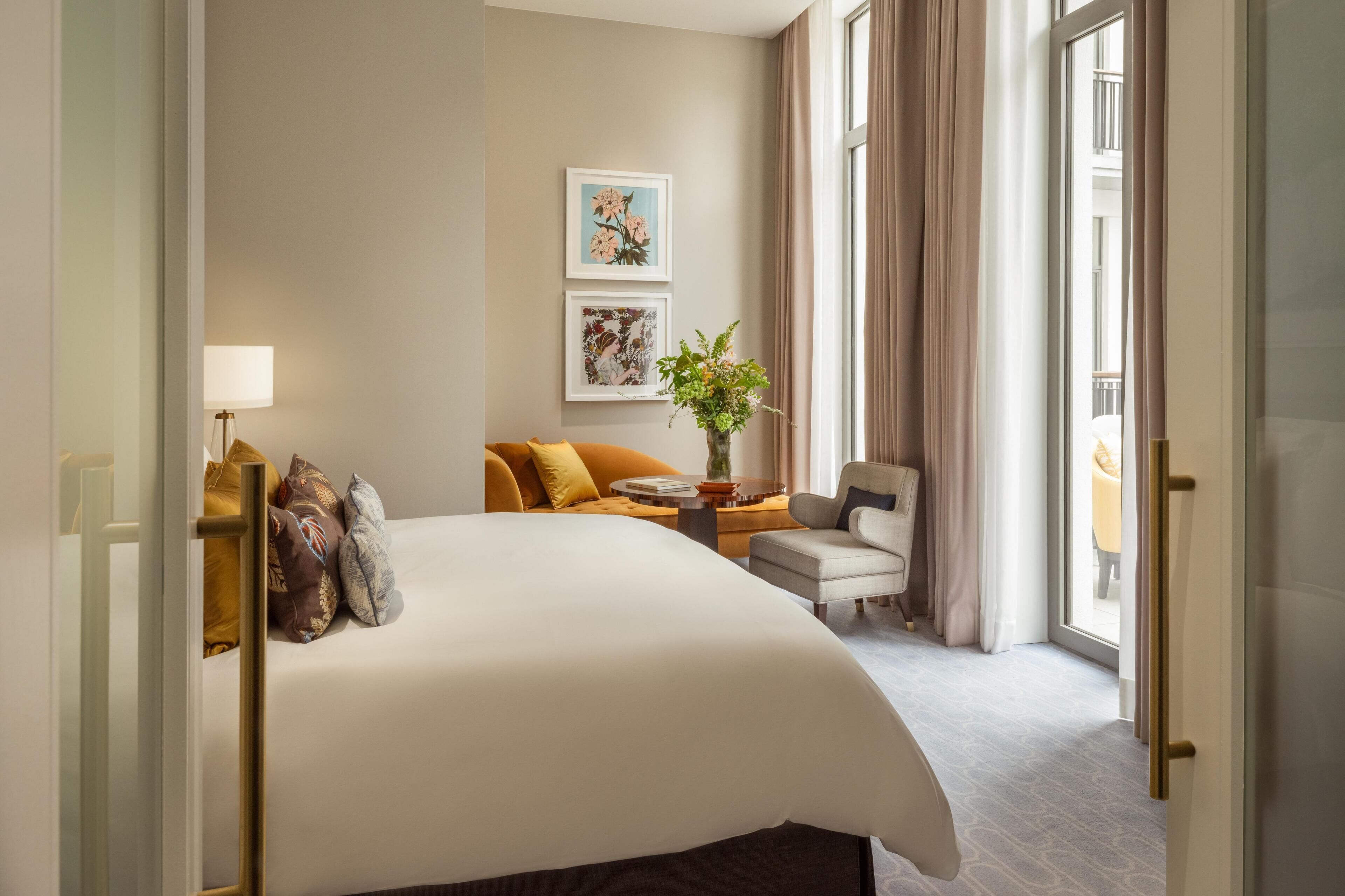 A hotel room with a large bed, a sofa, a chair, a small table, and a potted plant.