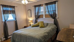 A bedroom with a bed, two windows with curtains, a nightstand with a lamp, and a wooden chair.