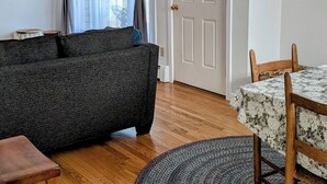 A living room with a sofa, a dining table with chairs, a wooden floor, and a door with curtains.