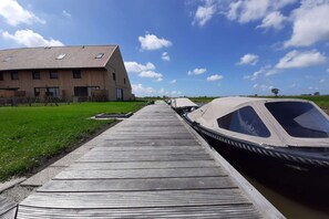 Un muelle de madera que lleva a un barco, un edificio con techo inclinado y un campo de césped bajo un cielo parcialmente nublado.