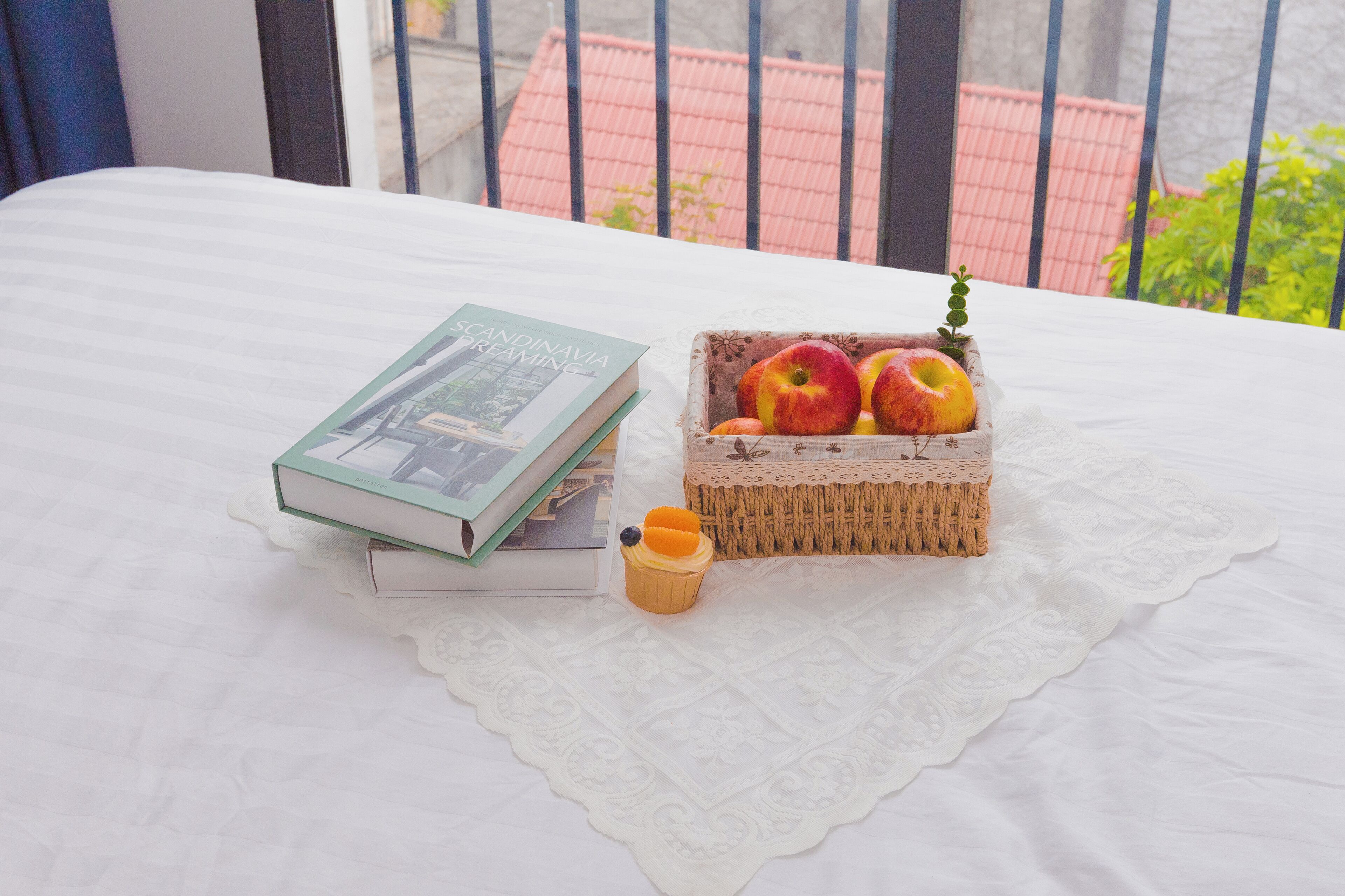 Uma cama bem arrumada com um bordado de renda, uma pilha de livros, uma cesta de vime com maçãs e uma xícara pequena.