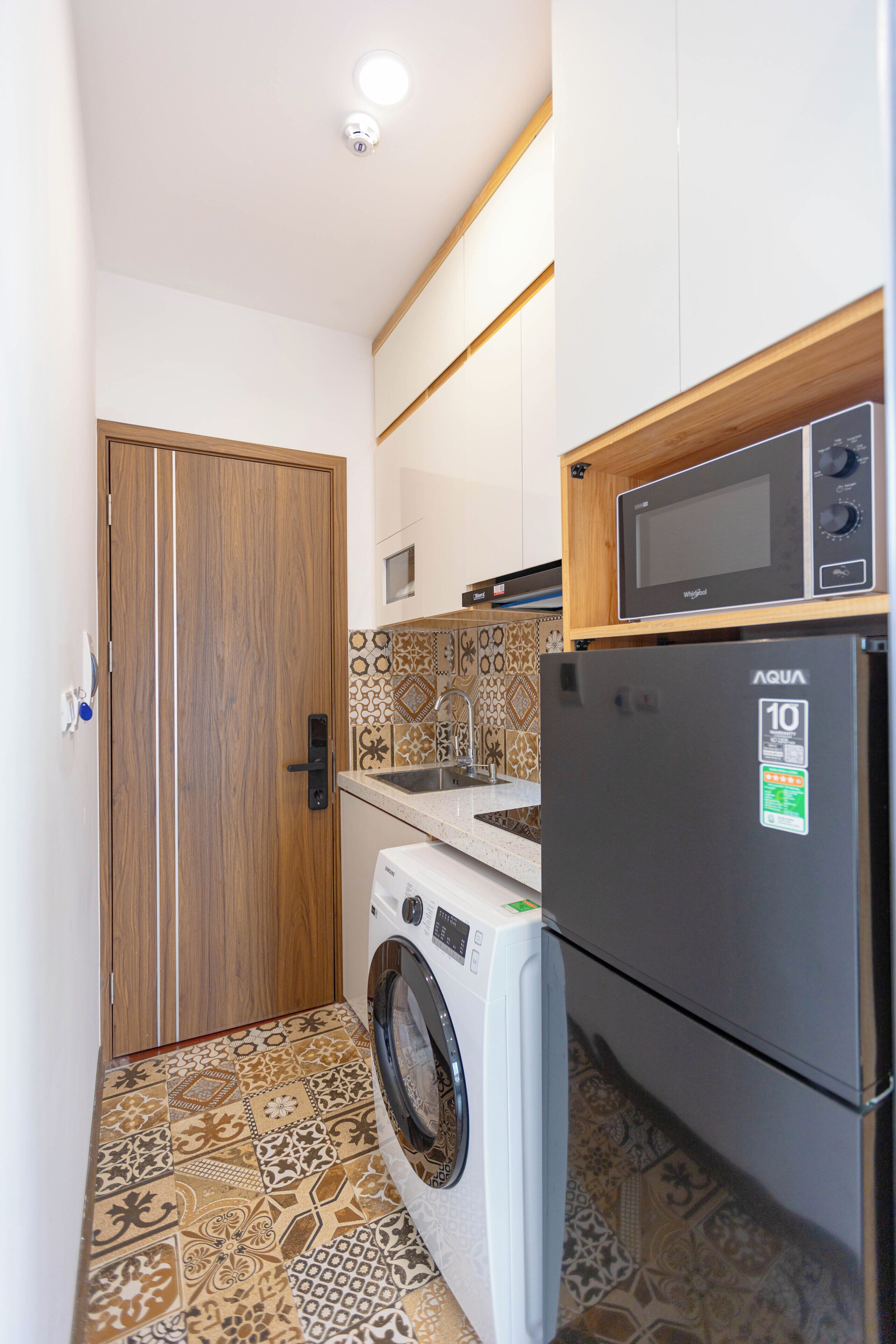 A compact kitchen with a washing machine, a refrigerator, and a microwave on a wooden shelf.