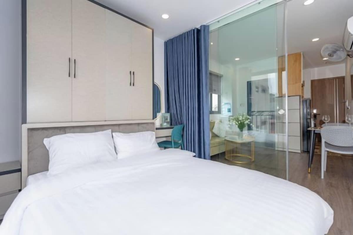 A modern hotel room with a large bed, a wardrobe, a desk with a chair, a glass partition, and a view of the kitchen area.