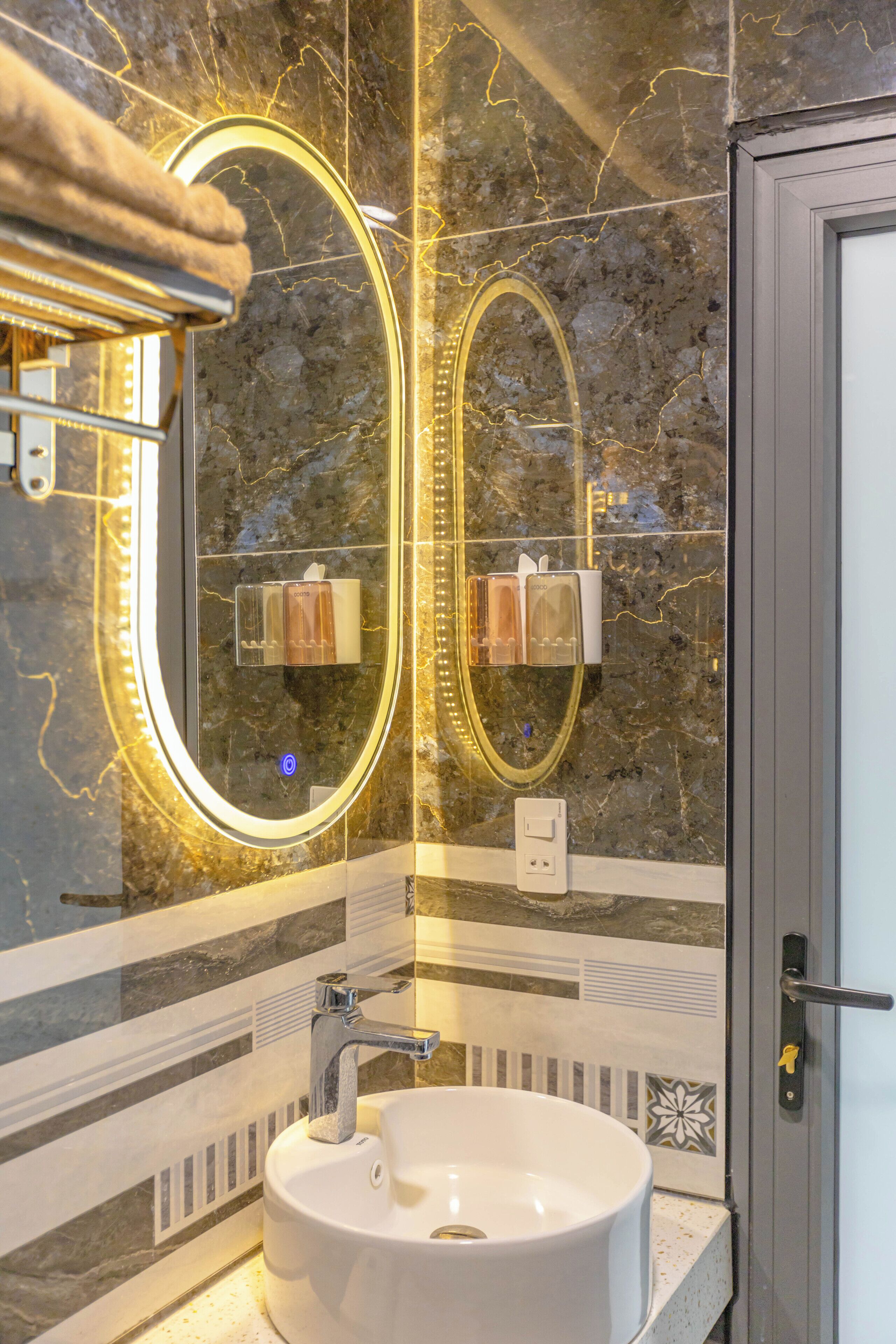 Une salle de bain avec un lavabo blanc, deux miroirs dotés de cadres éclairés, un porte-serviettes et un mur en marbre.