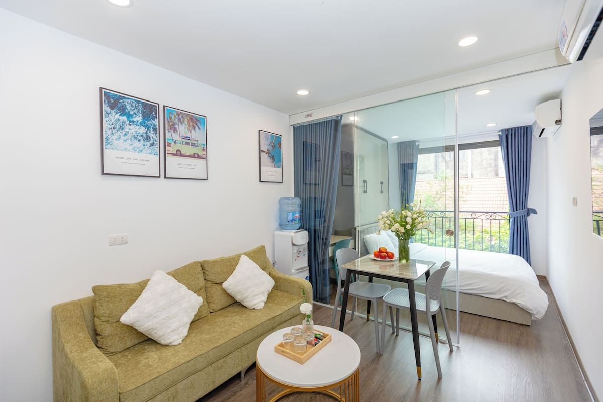 A modern hotel room with a sofa, a small table with a tray of snacks, a glass partition, a bed, and a view of the outdoors.