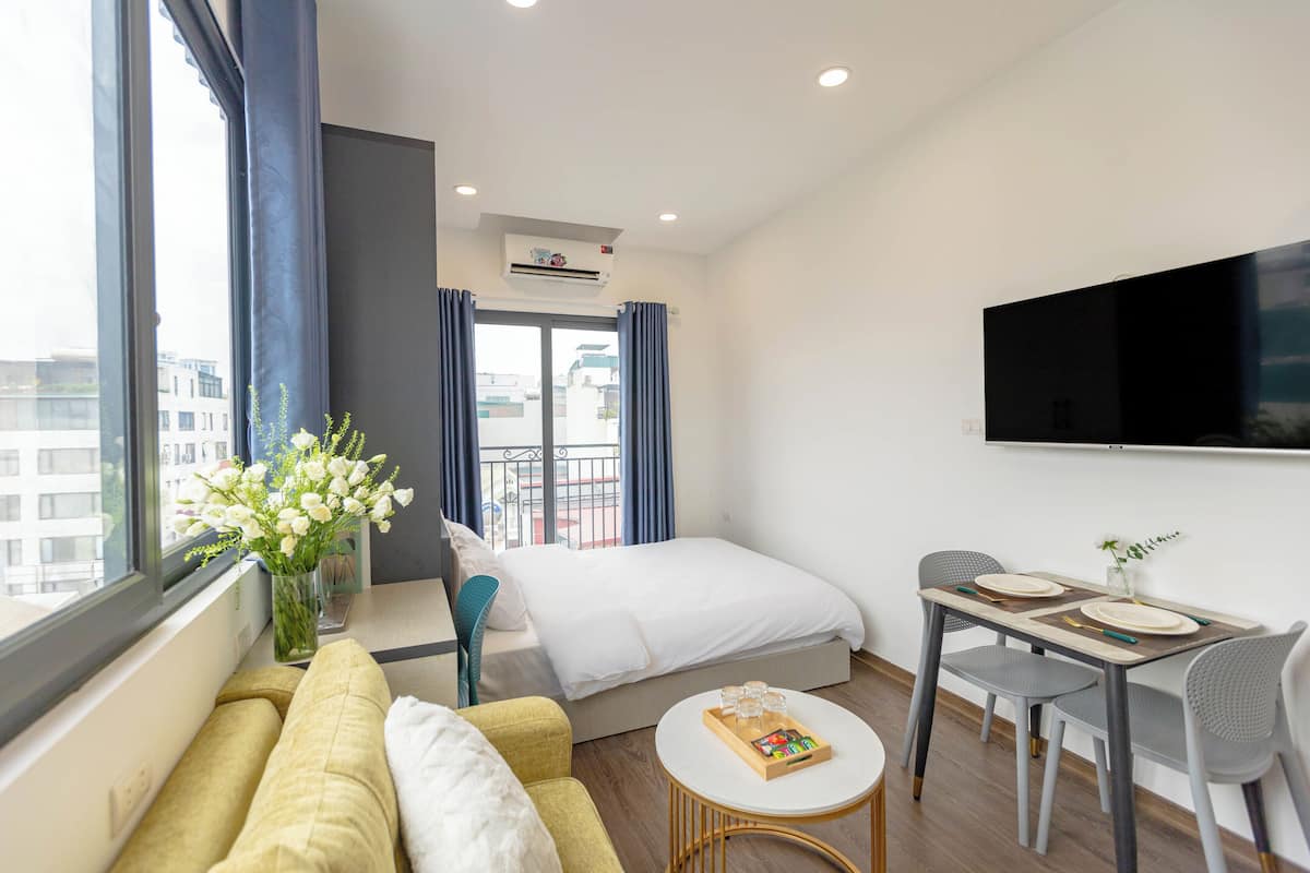 A modern hotel room with a bed, a sofa, a dining table, and a TV.
