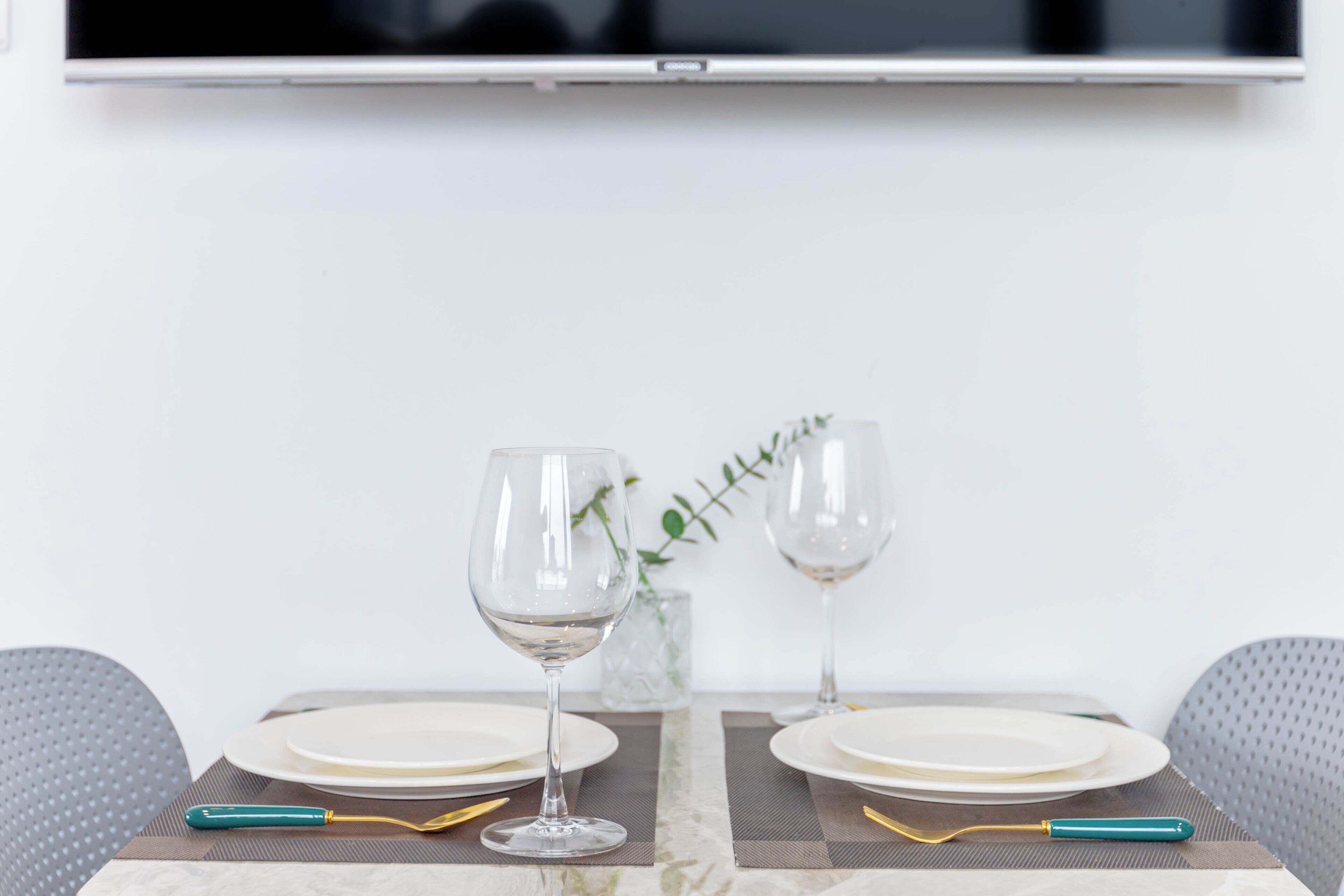 Una mesa de comedor dispuesta con platos, vasos y cubiertos, con una planta y un televisor arriba.