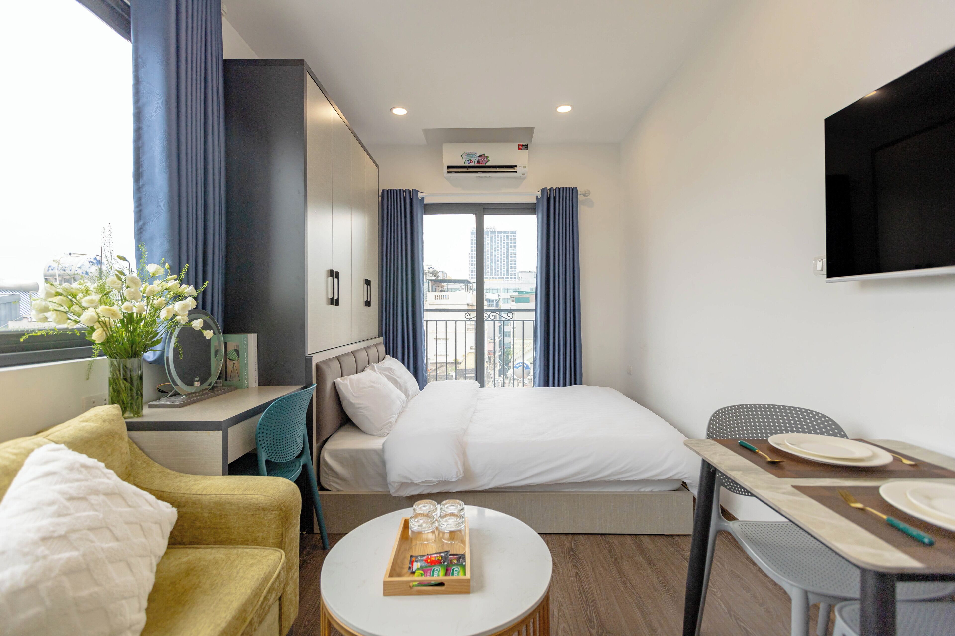 Habitación de hotel con cama, escritorio, silla, mesa de comedor, televisor y ventana con cortinas.