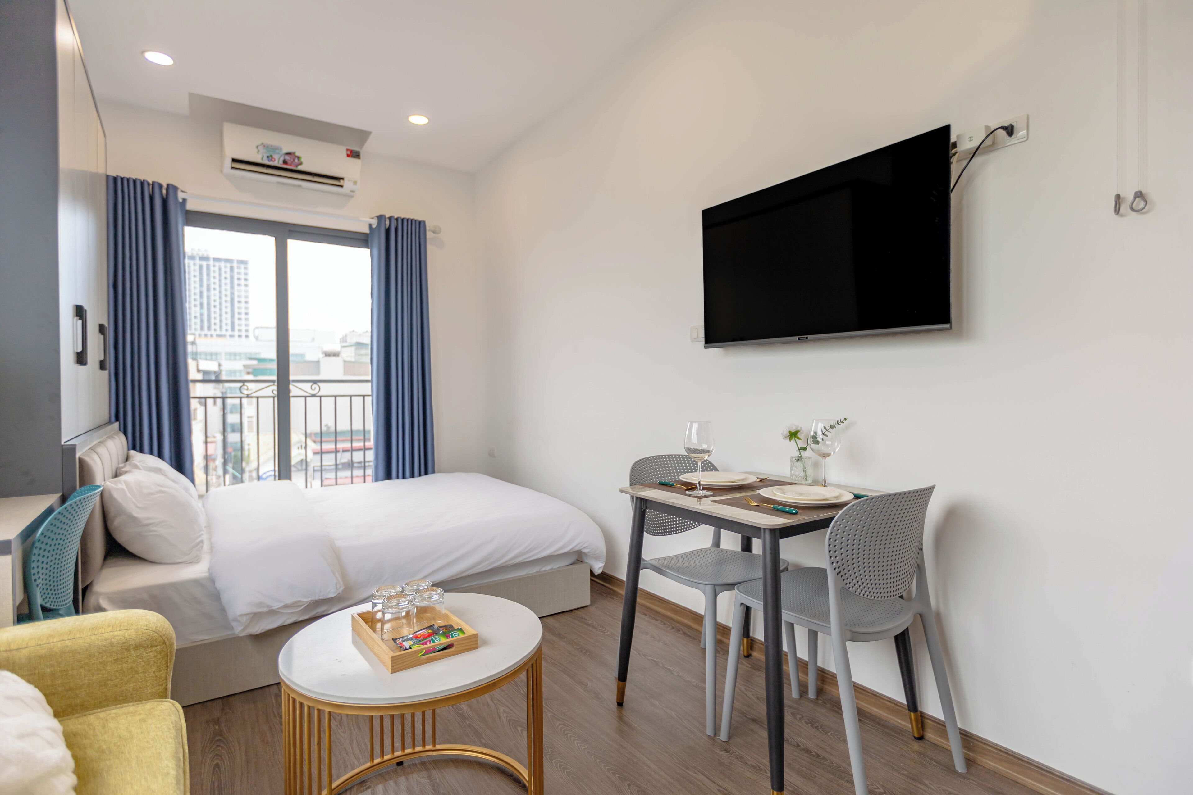 Una habitación de hotel con cama, una mesa de comedor con sillas, un televisor montado en la pared y una ventana con cortinas.