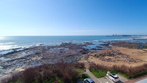 Une vue sur la côte, avec une plage rocheuse, un ciel dégagé et une silhouette de ville qui se dessine à l’horizon.