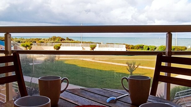 Una mesa de madera con cuatro tazas y un plato rojo con la palabra "VENICE", ubicada en un balcón con vistas a una zona verde y una masa de agua.