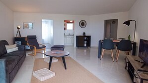 Una sala de estar con un sofá, dos sillones, una mesa de centro y un televisor. Se observa una cocina al fondo con armarios y un lavamanos.