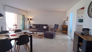 Una sala de estar moderna con un sofá, sillas, una mesa de comedor para dos personas y una zona de cocina con armarios y un reloj.