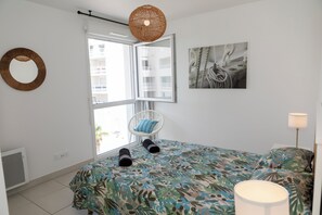 A bedroom with a bed, a chair, a hanging light, a window, and a wall-mounted mirror. - MY Hôtel Résidences - Port Leucate  (Leucate)