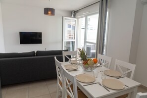 A modern living room with a sofa, a dining table set for two, and a flat-screen TV mounted on the wall. - MY Hôtel Résidences - Port Leucate  (Leucate)
