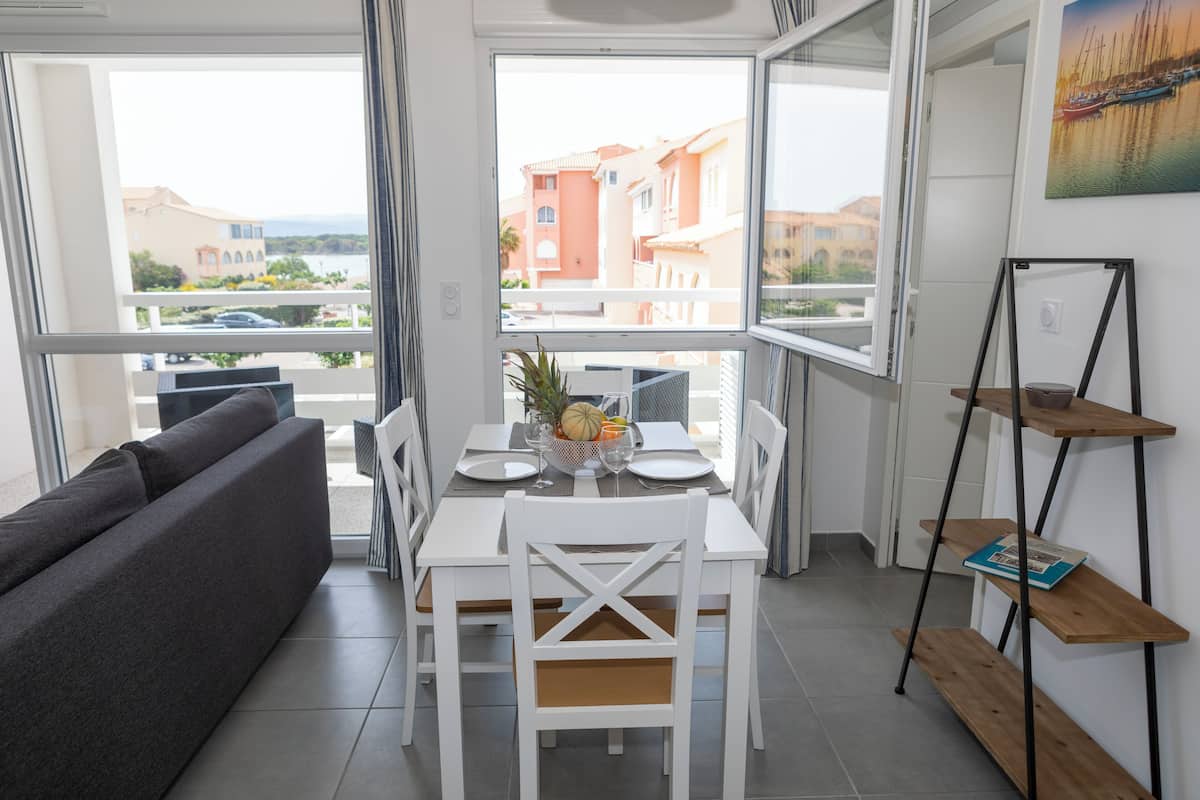 Ein moderner Essbereich mit einem weißen Tisch für zwei Personen, einer grauen Couch und Blick auf Gebäude durch große Fenster.