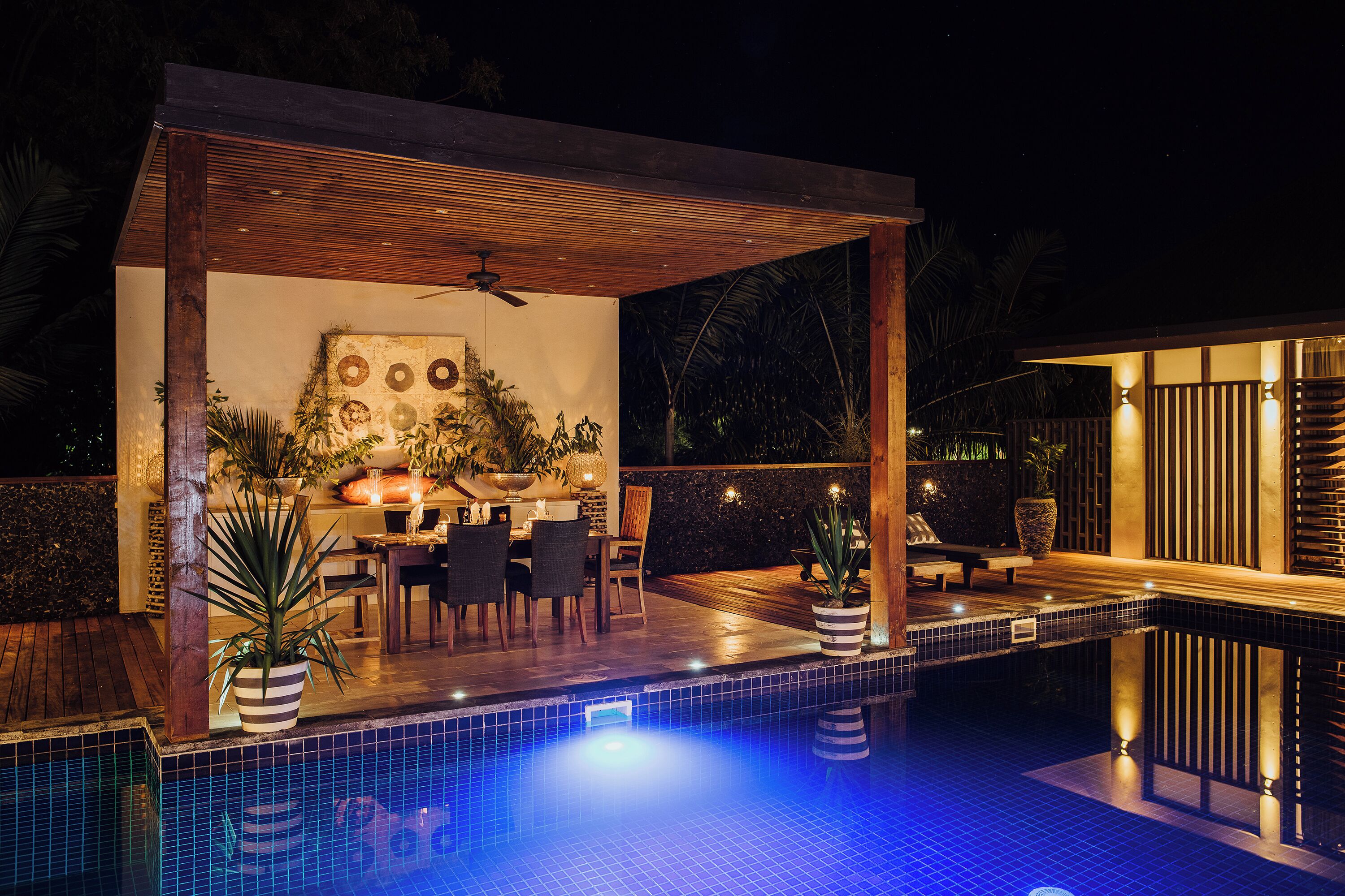 A poolside area with a covered patio, dining table, chairs, and potted plants.