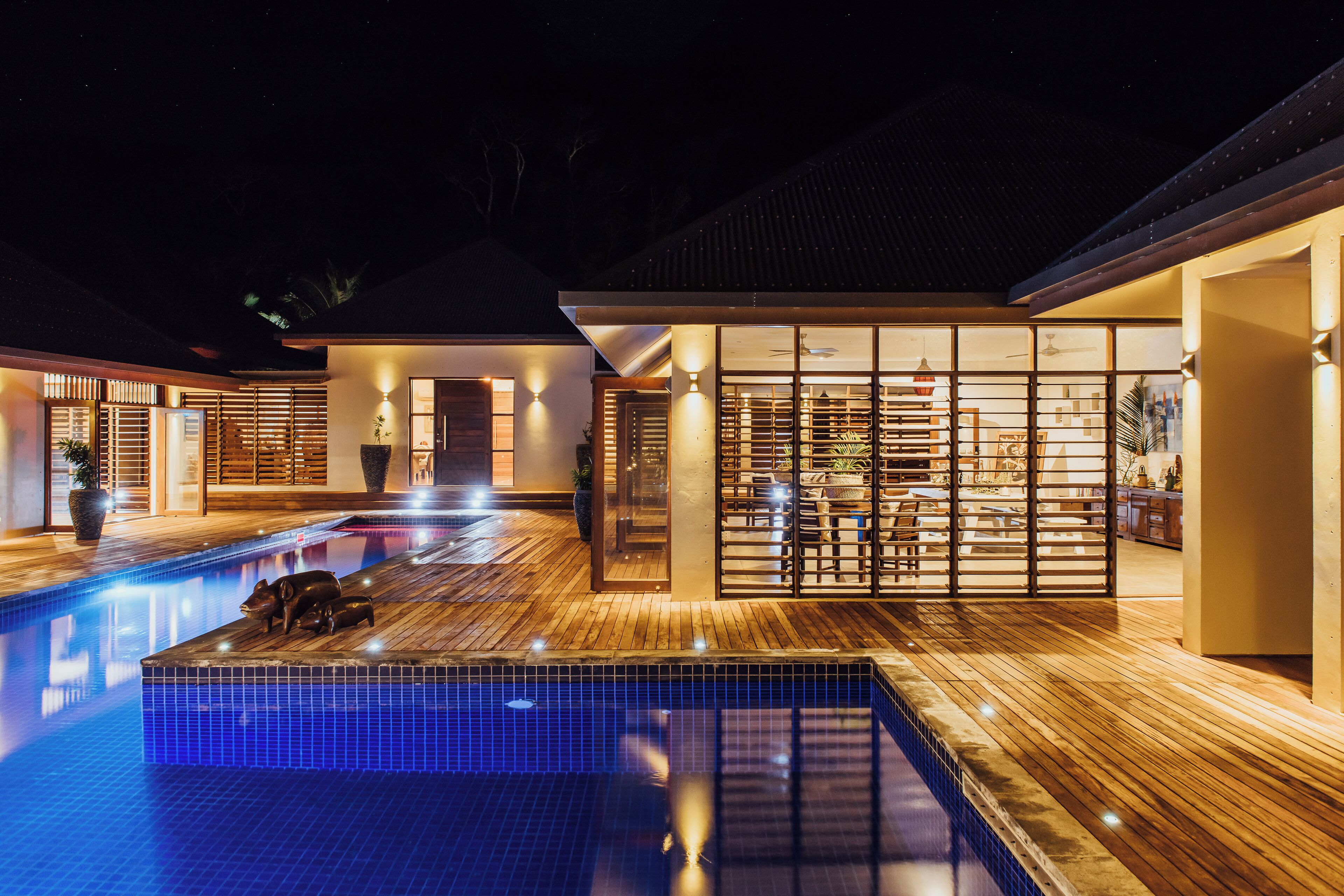 Un área de piscina moderna con terraza de madera, un espacio de estar con paredes de cristal y un interior bien iluminado.