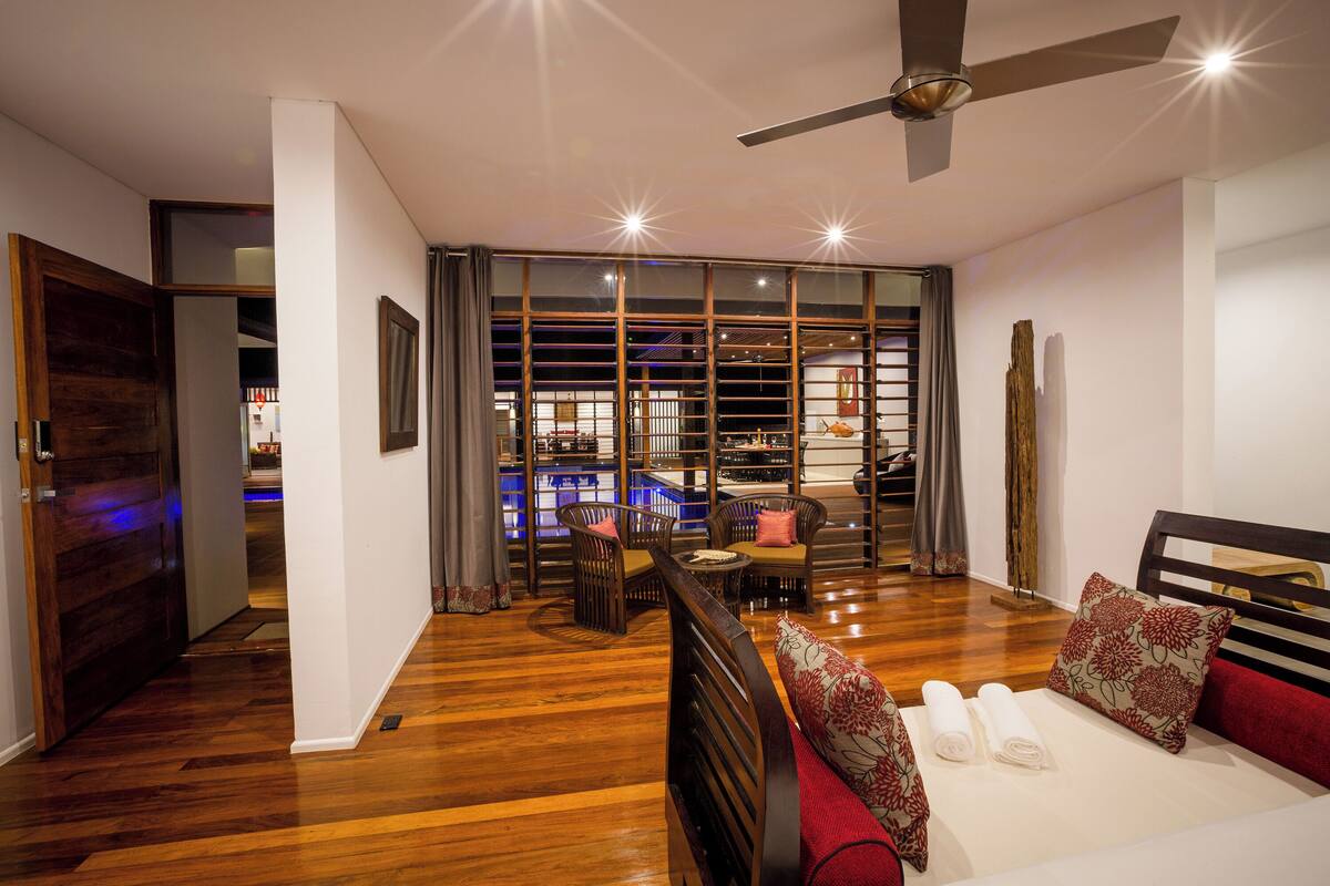 Une chambre d’hôtel avec un plancher en bois, un lit avec une literie rouge, un ventilateur de plafond et une vue sur un bar, visible à travers de grandes fenêtres.