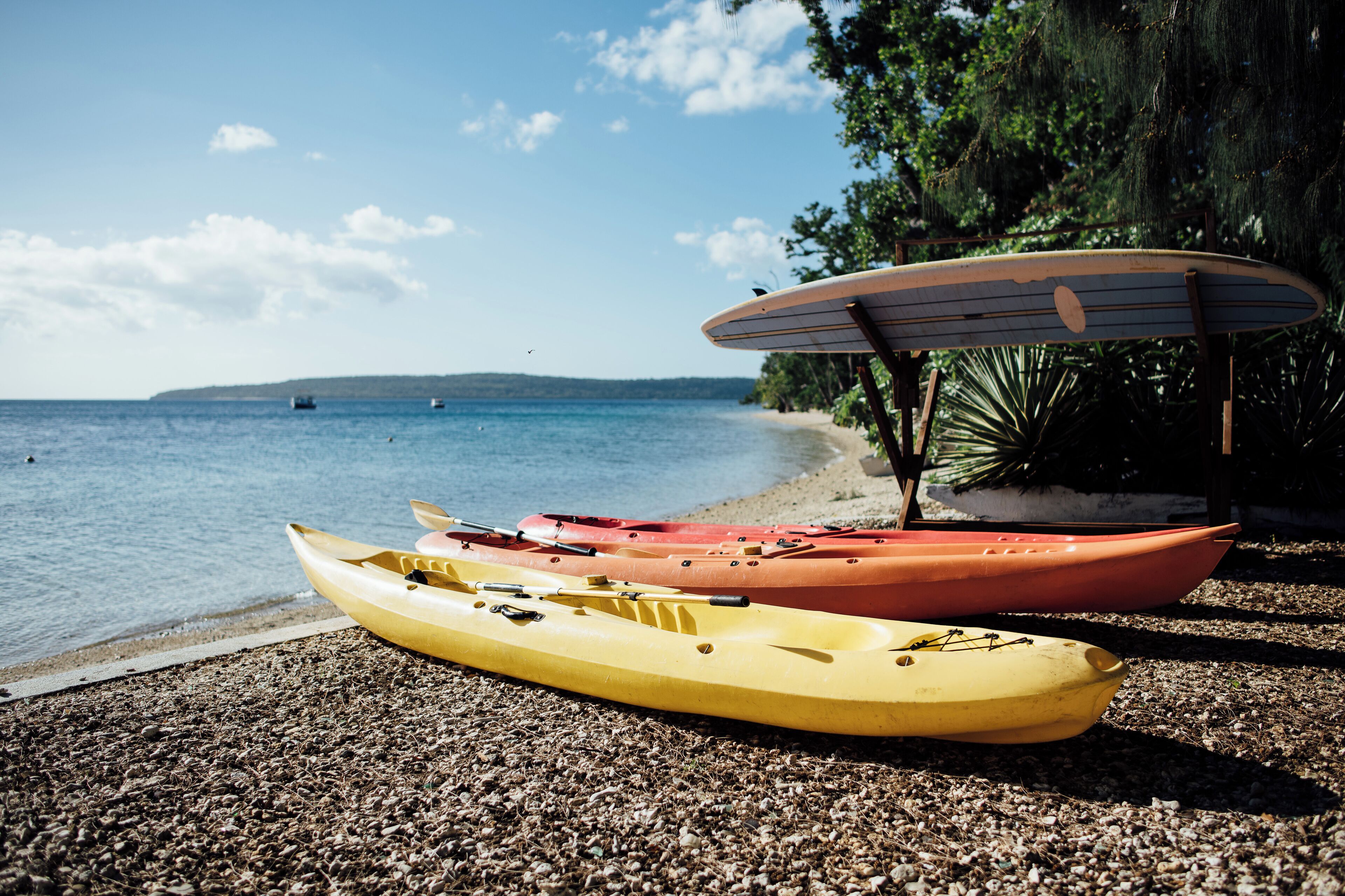 Kayaks y una tabla de surf en una playa de guijarros con mar en calma y una costa distante.
