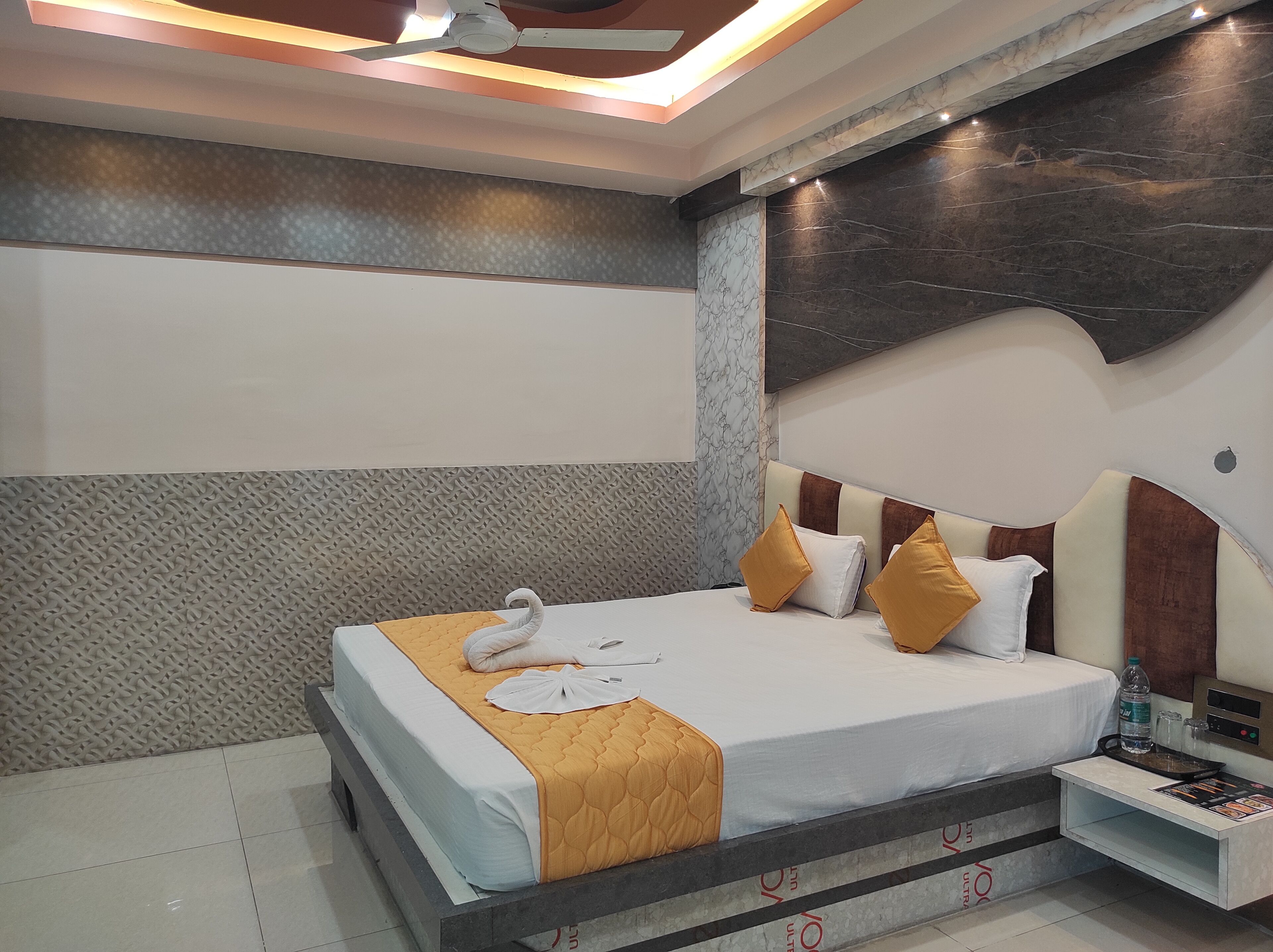 Una habitación de hotel con cama, mesita de noche, botella de agua y ventilador de techo.