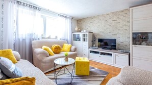 En moderne stue med sofa, lænestol, skærmpude, fjernsyn og en murstensvæg.