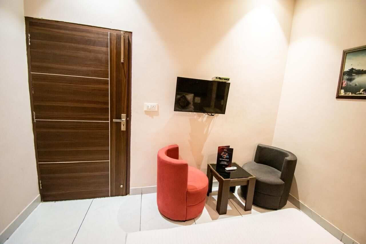 Una habitación con puerta de madera, un televisor de pantalla plana en la pared, una silla roja, un sillón gris y una pequeña mesa con un control remoto.