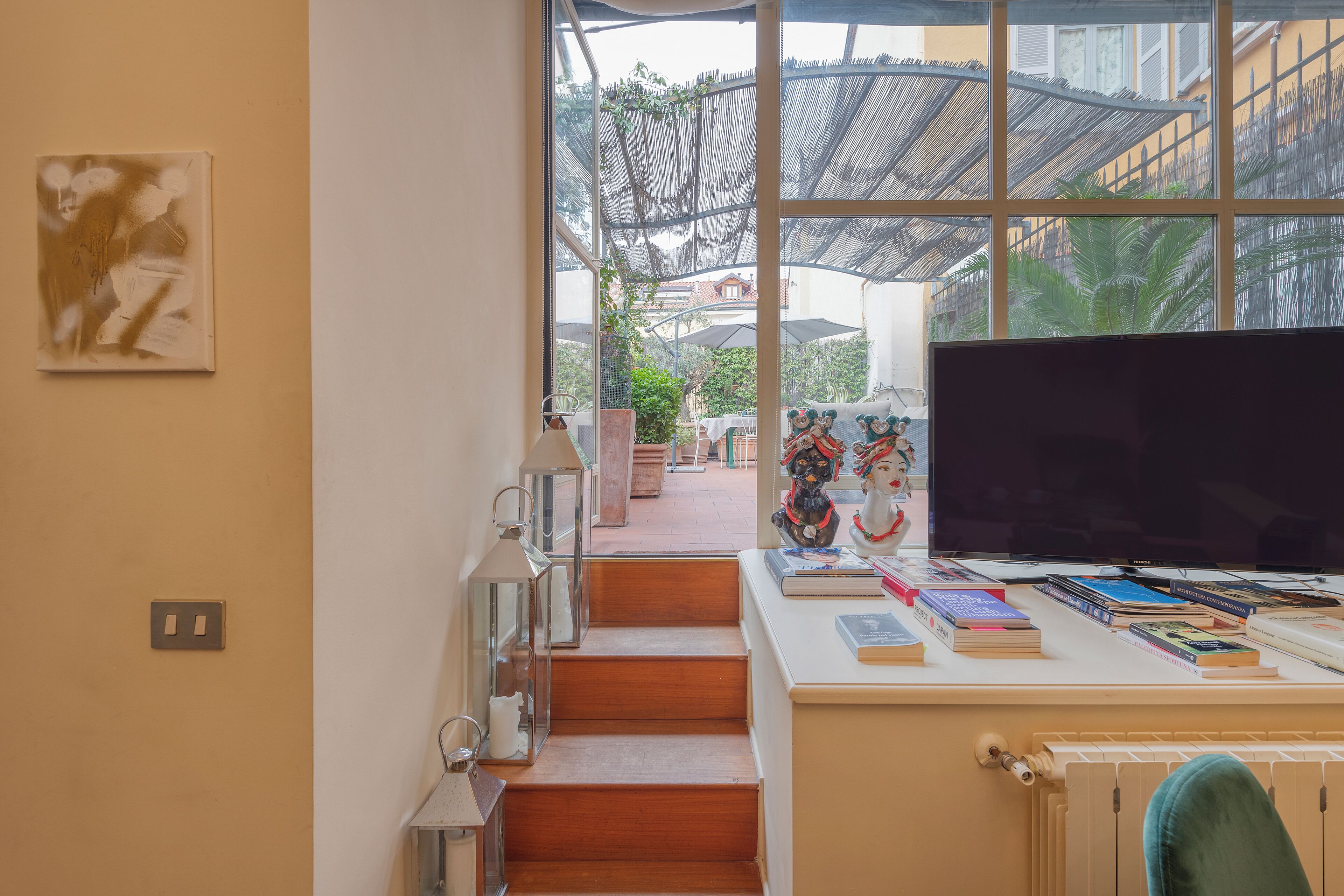 Una habitación con un gran ventanal, un escritorio con libros y un televisor, y una escalera que lleva a un espacio al aire libre.