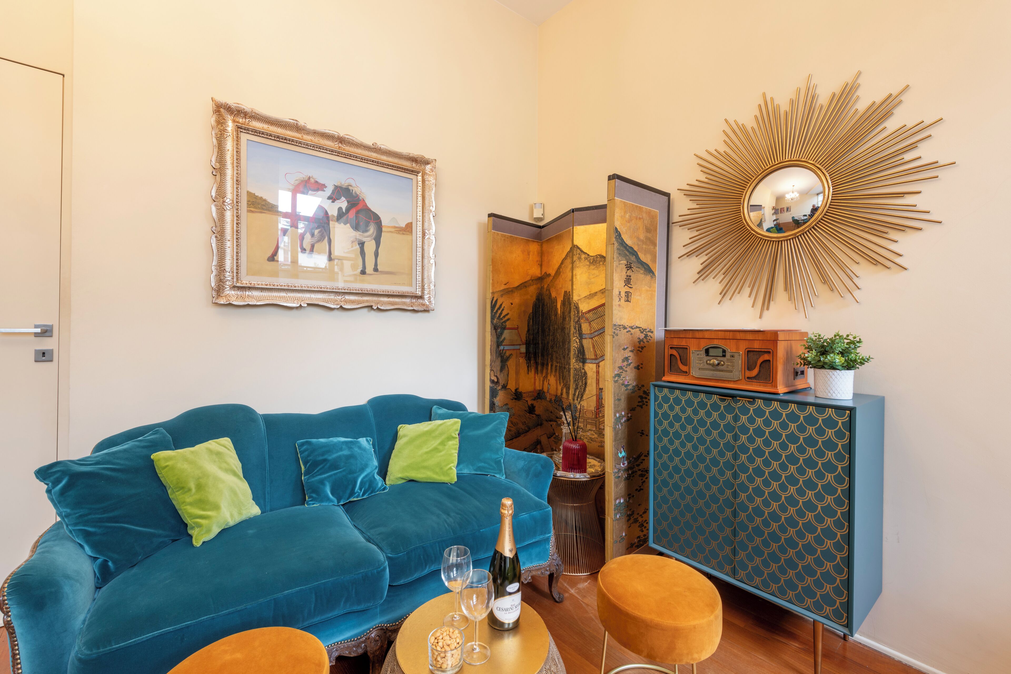 A living room with a blue sofa, a small round table with a bottle and glasses, a decorative mirror, and a cabinet with a patterned door.