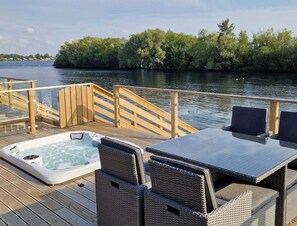 A wooden deck with a hot tub, wicker chairs, and a table overlooking a calm body of water and greenery. - Rolo's Dog Friendly Lakeside Lodge With Hot Tub (Stamford)
