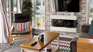Un saló amb una paret de maó, un televisor pantalla plana muntat sobre una xemeneia, una taula de centre de fusta, un sofà i una cadira amb un coixí de colors.