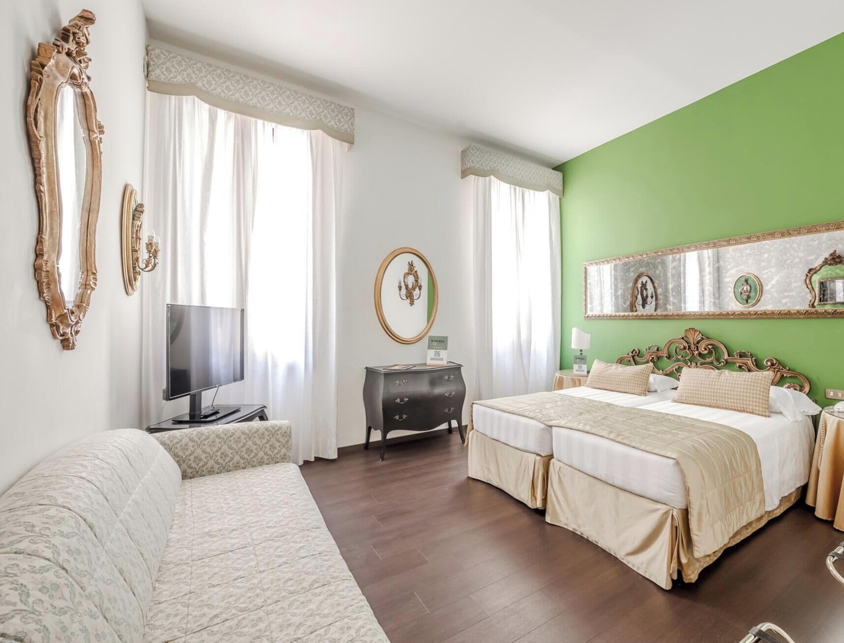 Quarto de hotel com cama de casal, sofá, televisão, penteadeira e parede verde decorativa.