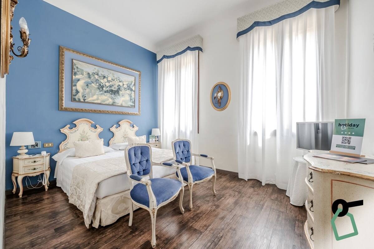 A bedroom with a bed, two chairs, a nightstand, a lamp, a painting, a decorative plate, and a sign on a desk.