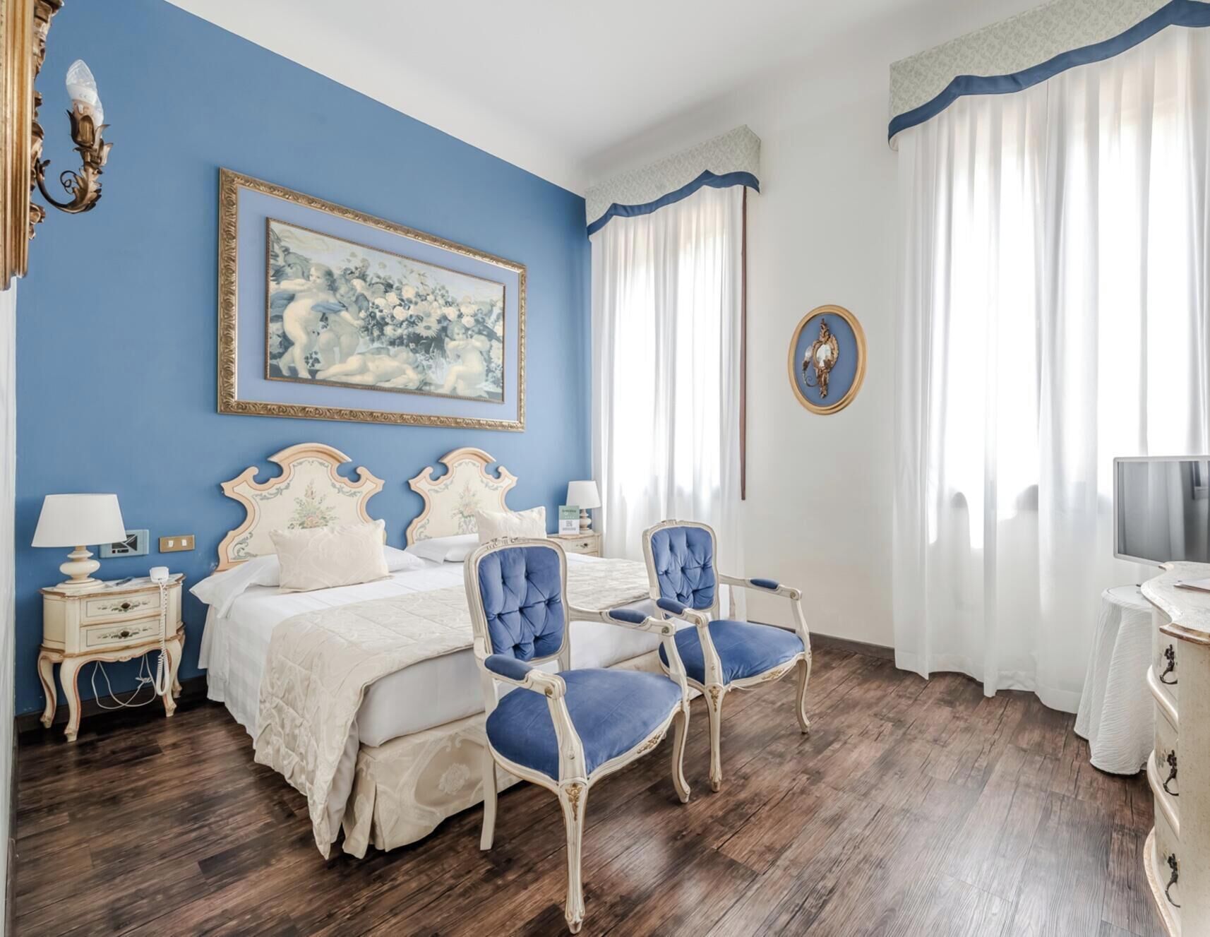 Ein Schlafzimmer mit einem Bett, zwei Stühlen, einem Nachttisch, einer Lampe, einem Gemälde, einem gerahmten Bild und einem Schreibtisch mit Computer und Schild.