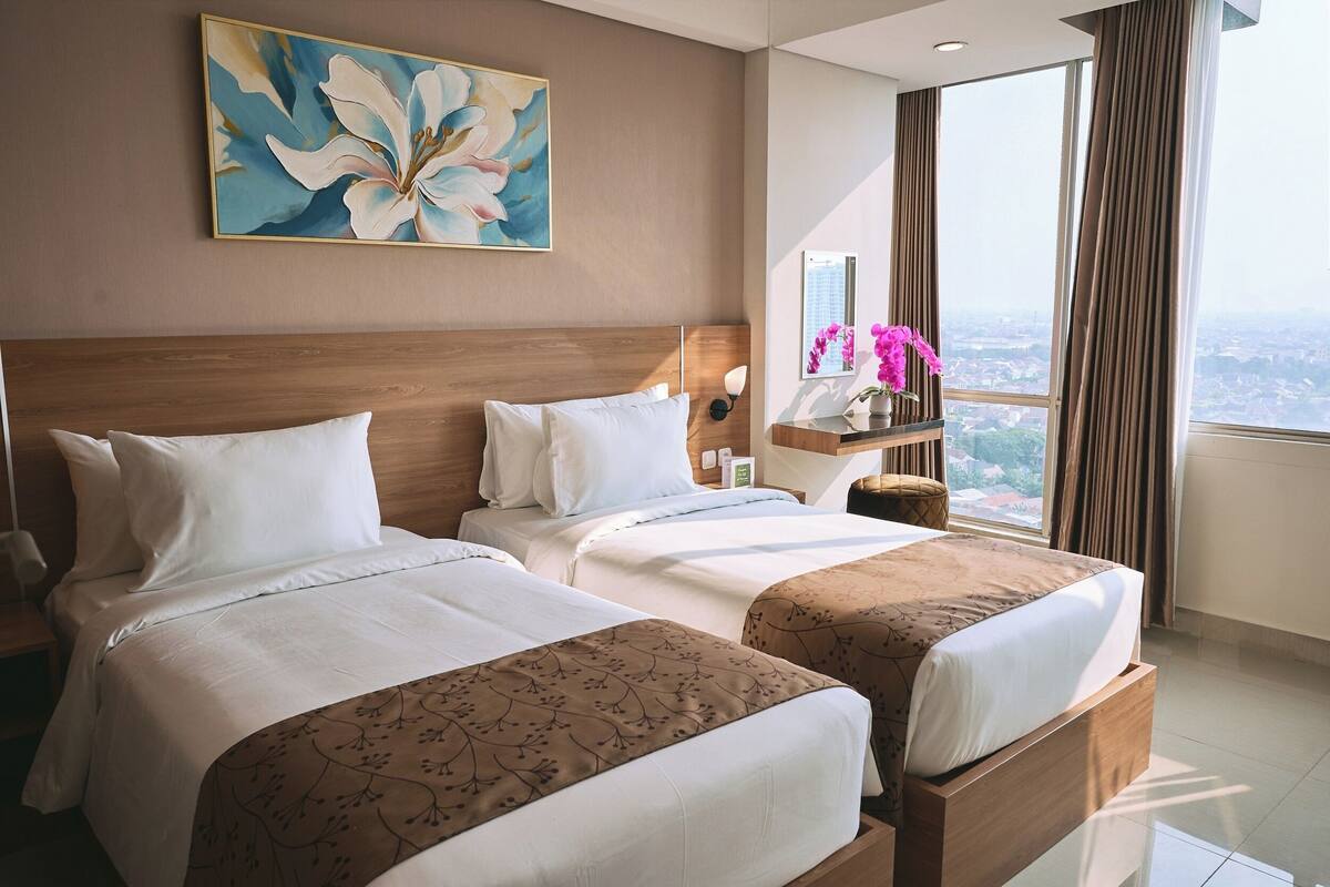 Habitación de hotel con dos camas, un ventanal con cortinas, un cuadro en la pared y una mesita de noche con lámpara.