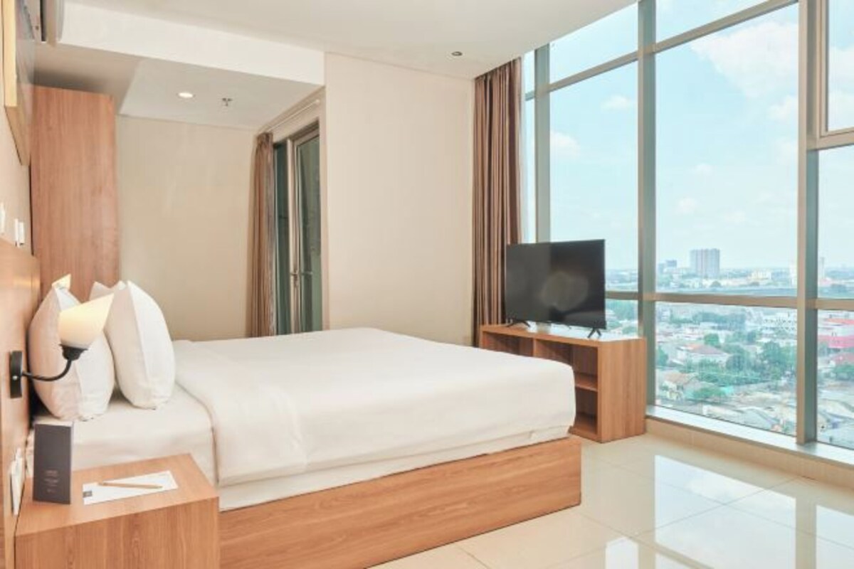 Une chambre d’hôtel moderne avec un grand lit, un téléviseur posé sur un meuble en bois, une table de chevet et une vue sur la ville grâce à de grandes fenêtres.