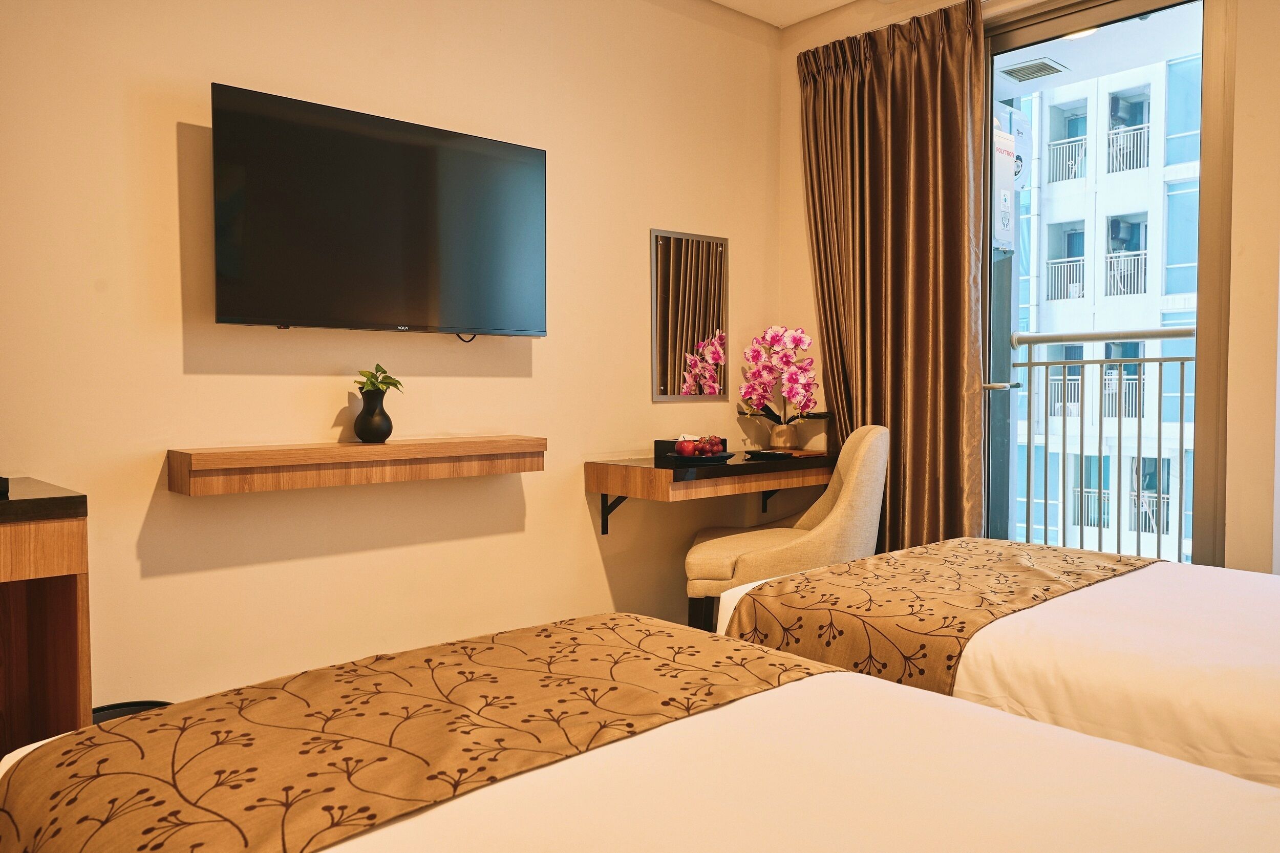 A hotel room with two beds, a TV, a desk with a chair, a vase with flowers, and a view of a building through the window.