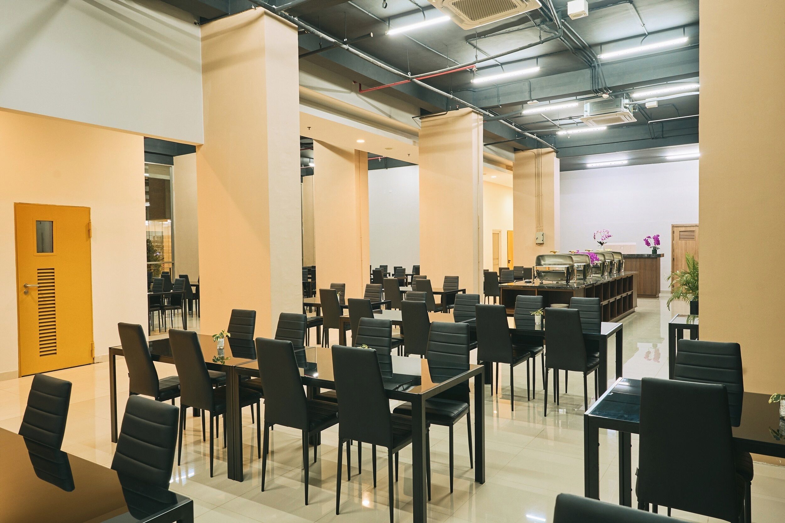 A spacious conference room with multiple tables and chairs, a reception area with a counter and plants, and a modern ceiling with exposed pipes and lighting.