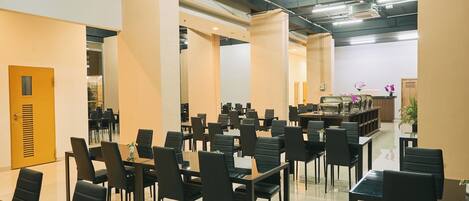 A spacious conference room with multiple tables and chairs, a reception area with a counter and plants, and a modern ceiling with exposed pipes and lighting.