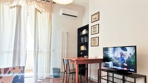 A living room with a TV, a dining table, chairs, a bookshelf, and a window with curtains. - Naples on foot (Napoli)