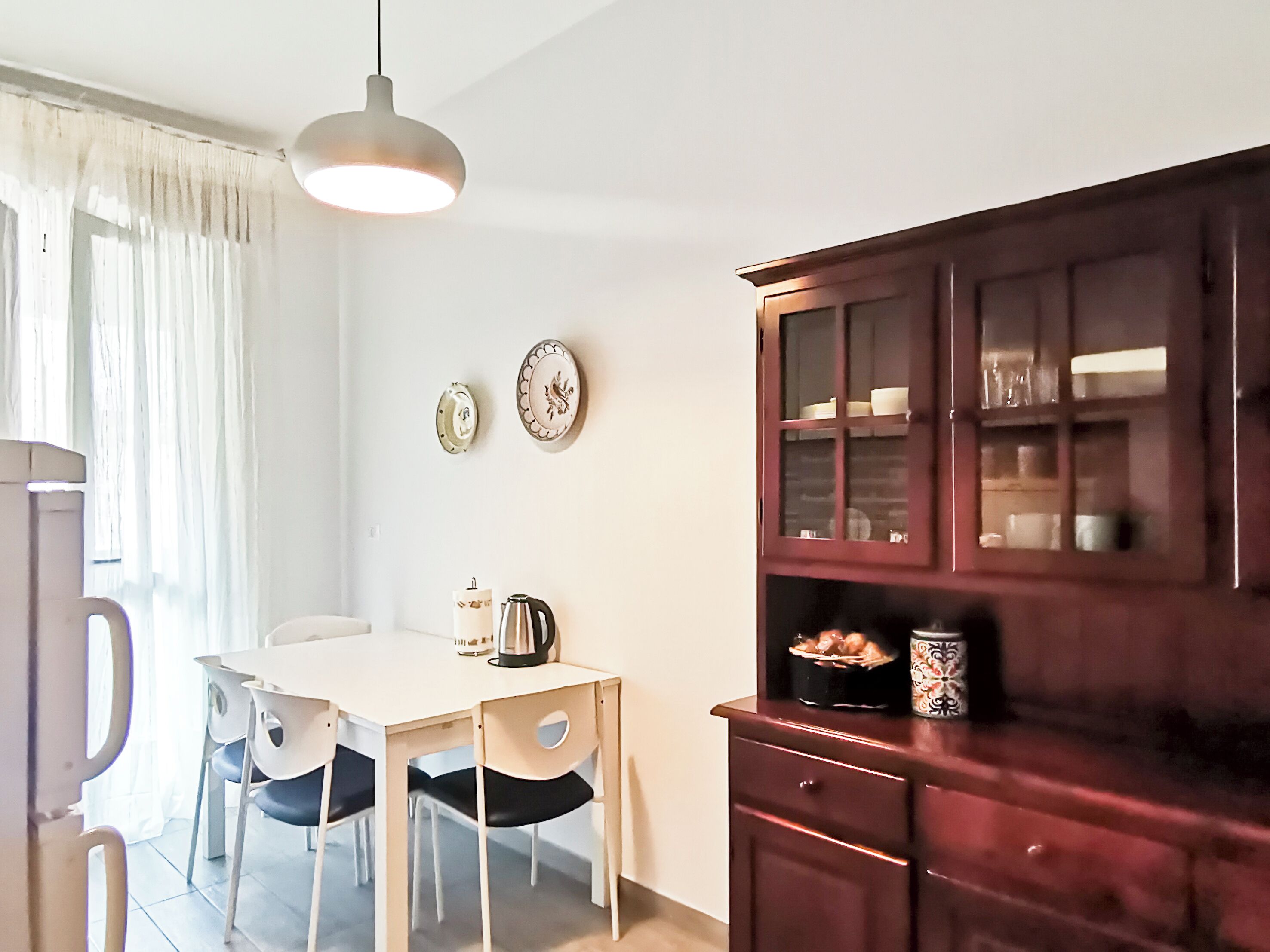 A dining area with a white table and chairs, a hanging light fixture, and a wooden cabinet with glass doors.