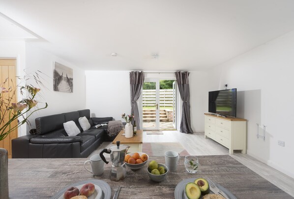 A modern living room with a black leather sofa, a coffee table with fruits and drinks, a TV, and a sliding glass door leading to an outdoor area.
