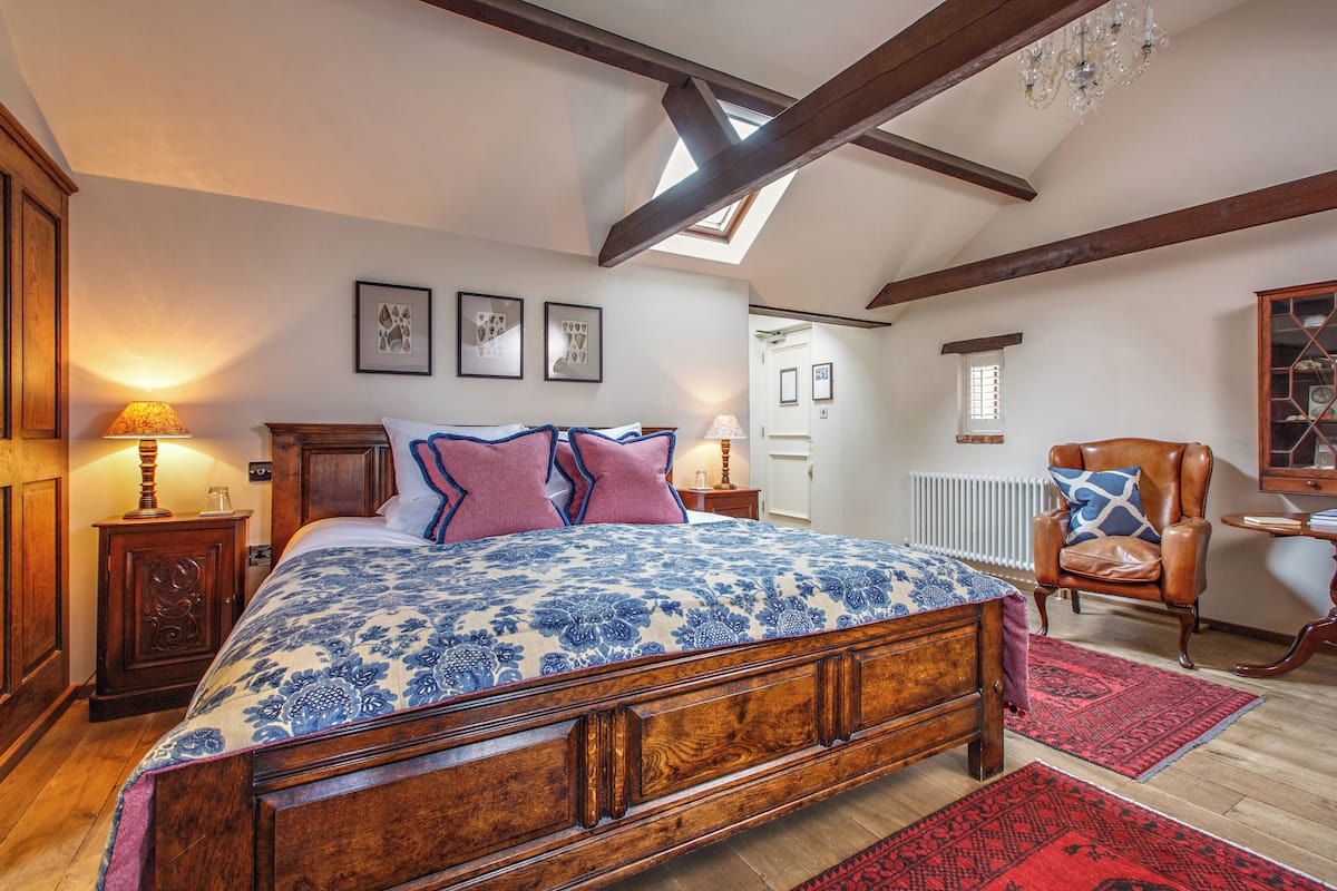 A bedroom with a wooden bed, a red and blue patterned bedspread, a wooden nightstand with a lamp, a wooden chair, and a red rug.