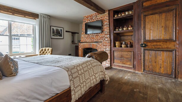 A bedroom with a large bed, a brick fireplace, a television mounted on the wall, and wooden shelves with decorative items. - Byfords  (Holt)
