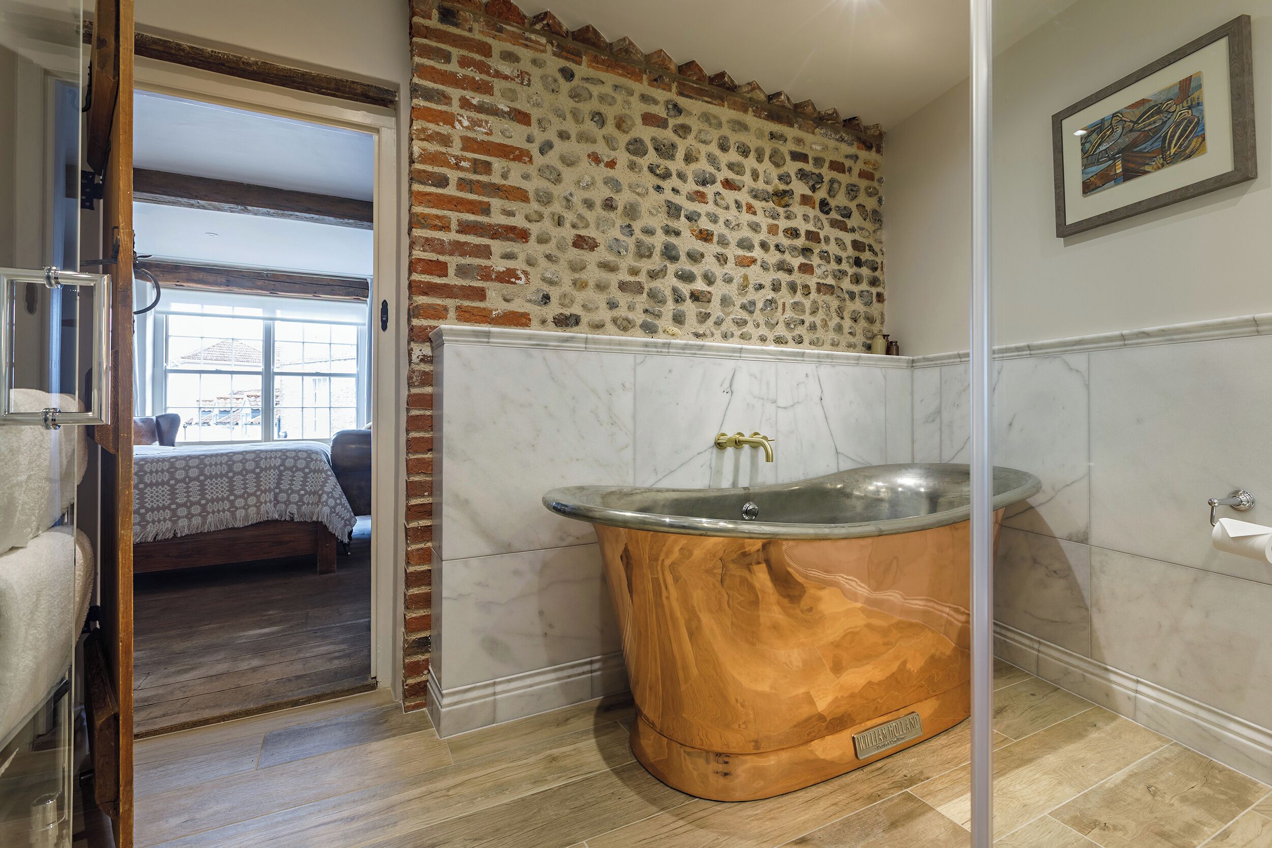 Un baño con una bañera de madera, una pared de piedra y una cama visible a través de una puerta abierta.