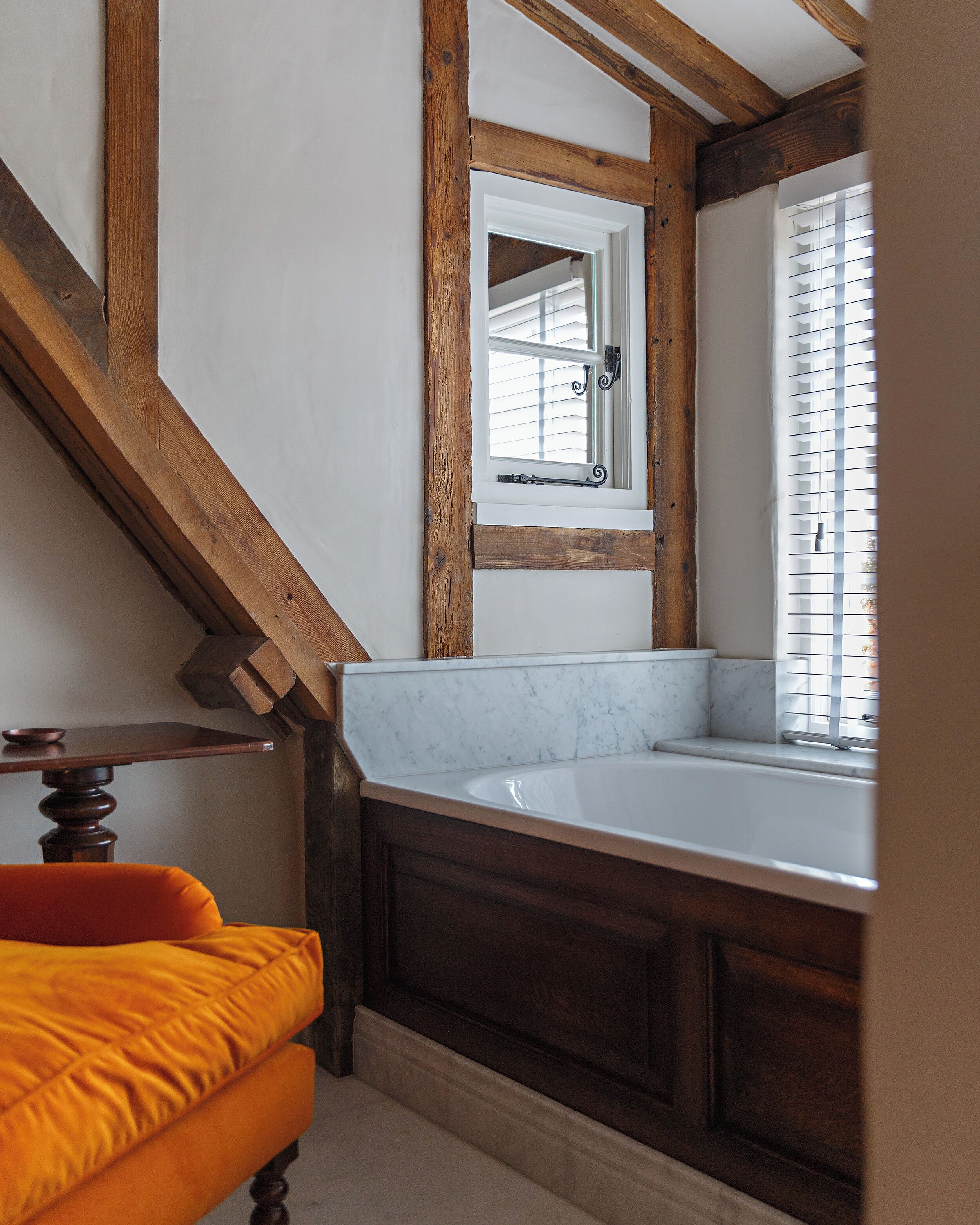 Une salle de bain avec une baignoire blanche, des poutres en bois, une fenêtre avec des stores et une chaise orange.