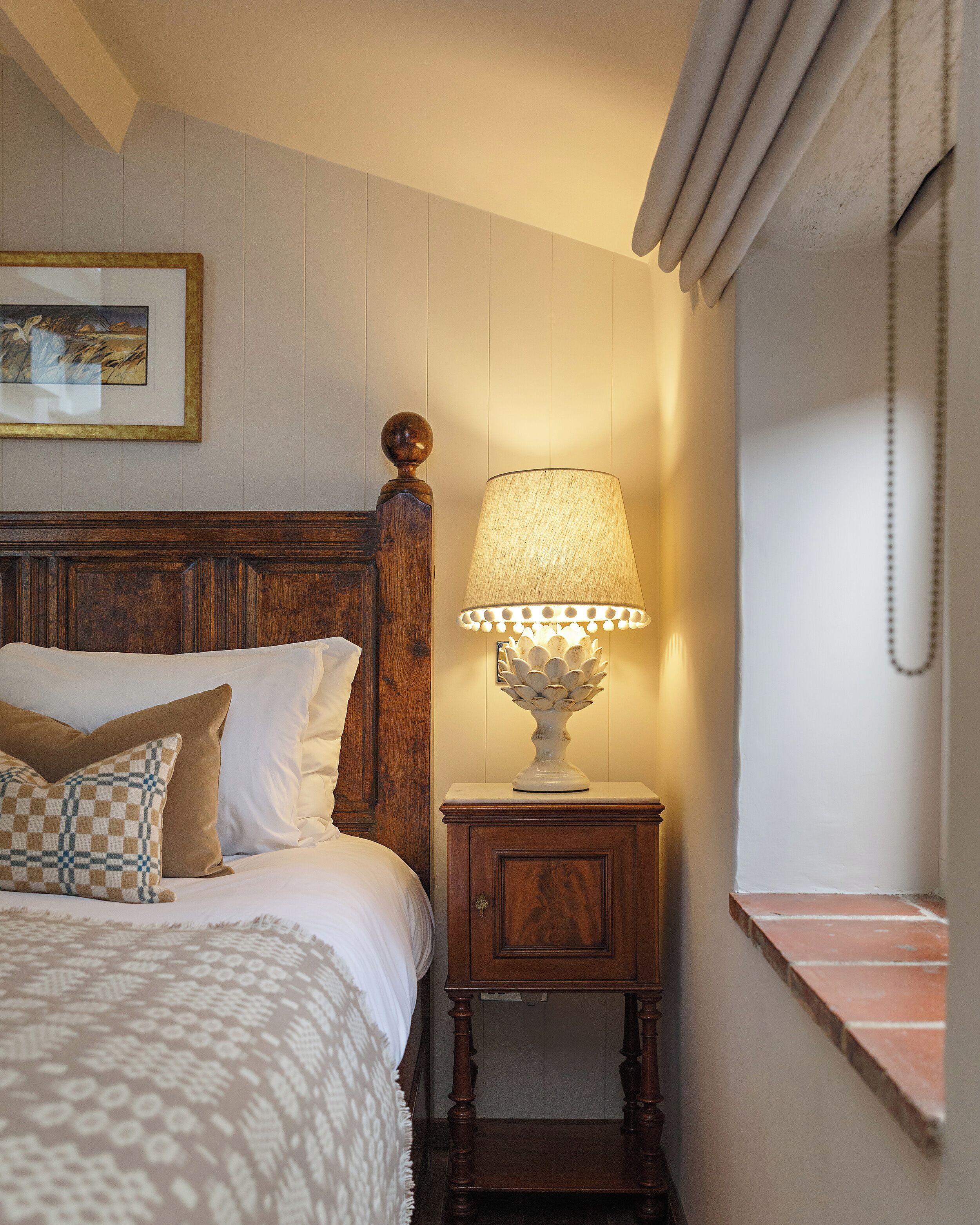 Een netjes opgemaakt bed met een houten hoofdbord, een nachtkastje met een lamp, en een ingelijste foto aan de muur.