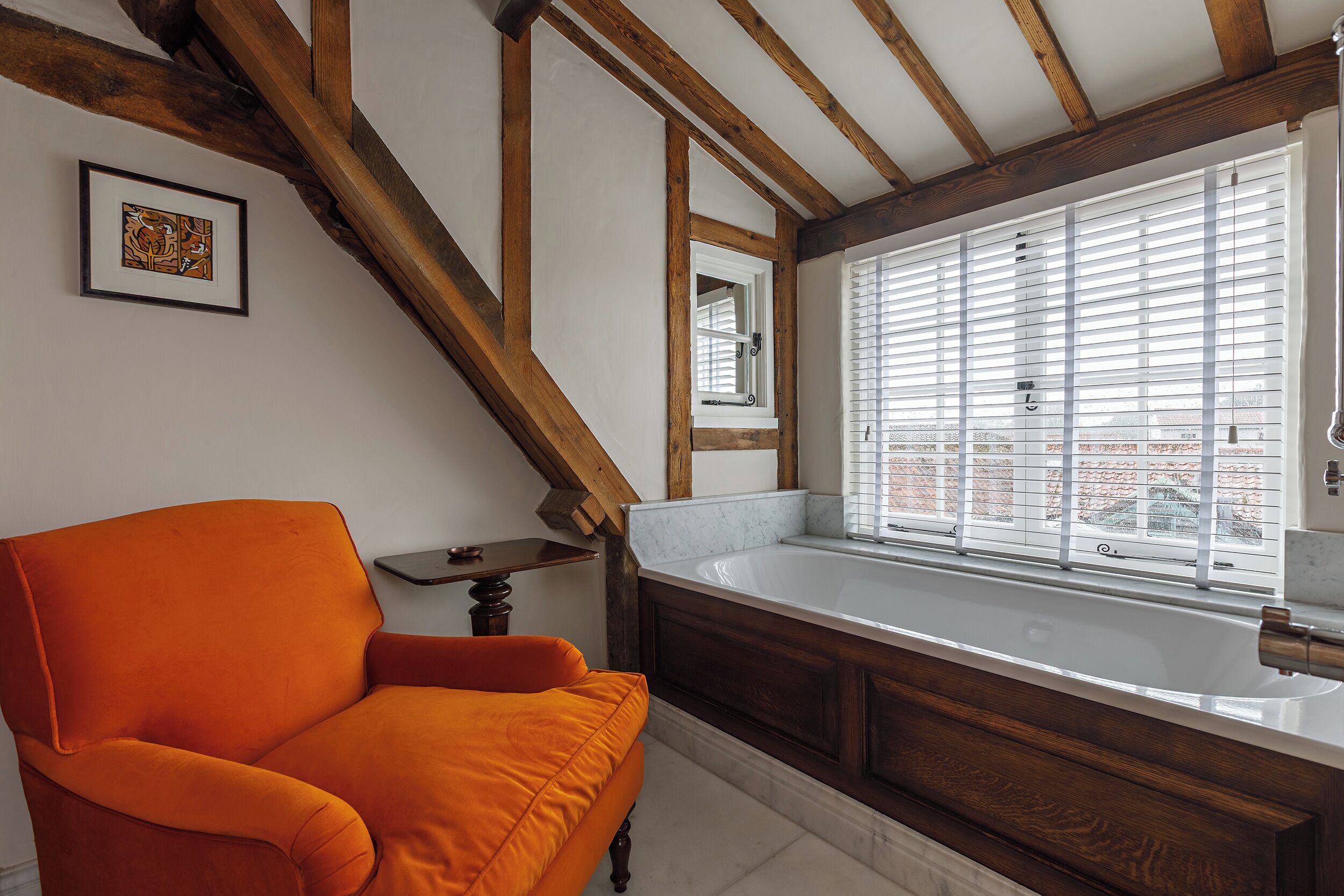 A room with a wooden ceiling, a large window with blinds, a white bathtub, and an orange armchair.