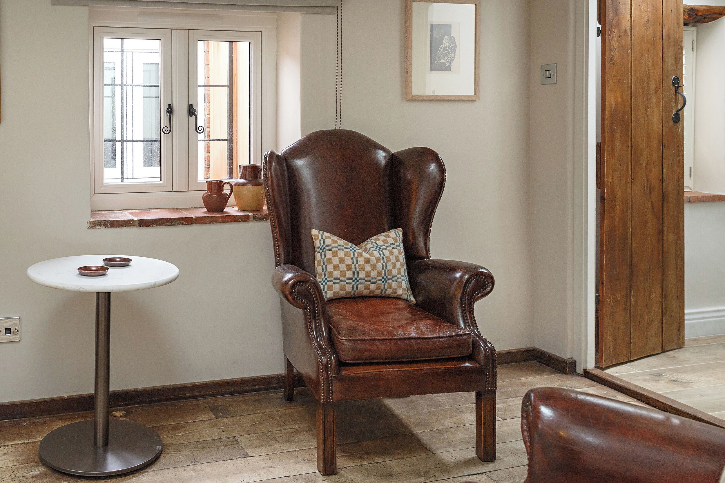 A room with a brown leather armchair, a small round table with two bowls, a window with a view, and a framed picture on the wall.